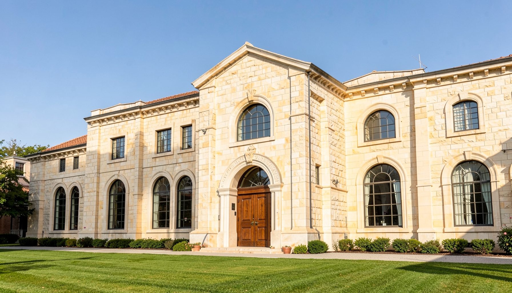 Sunlit stone institutional building facade with arched windows and a manicured lawn, warm beige and cream tones